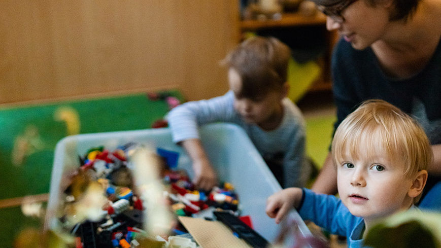 Im Vordergrund schaut ein Kind frontal in die Kamera. Im Hintergrund ein weiteres Kind Lego spielend. Eine Erzieherin kniet hinter den beiden lächelnd.