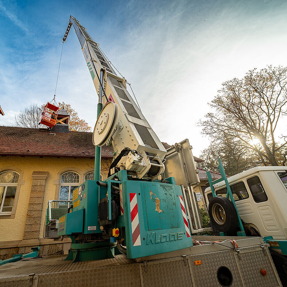 Zwei Dachdecker in einem Arbeitskorb, der an einem Kran hängt, reparieren den Klockenturm der Kirche des Epilepsiezentrums Kleinwachau