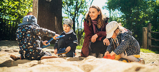 Eine Erzieherin sitzt mit drei Kindern im Sandkasten der Außenanlagen der Kita Schatzinsel in Pulsnitz in Sachsen. Die Kita Schatzinsel gehört zum Epilepsiezentrum Kleinwachau in Radeberg.