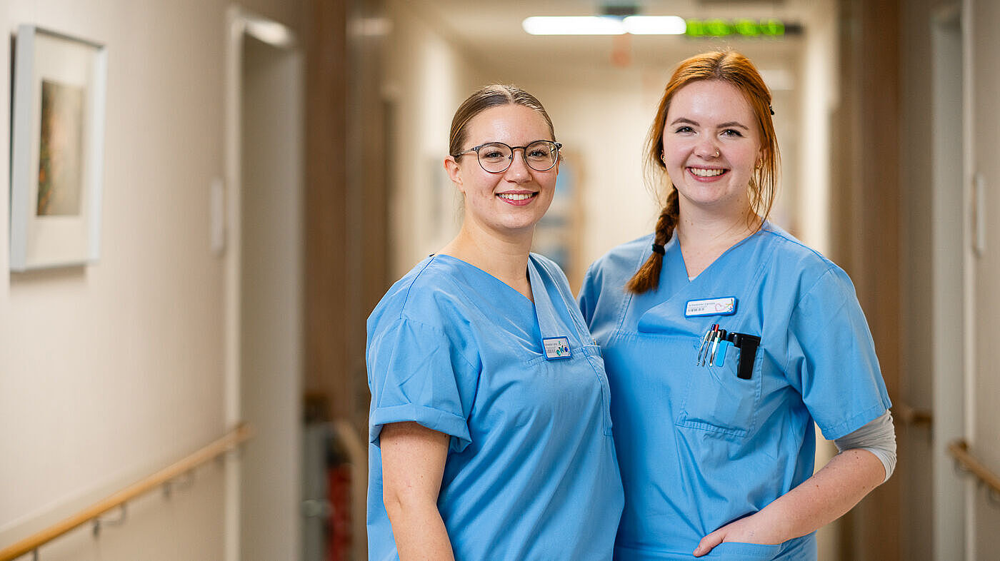 Schwester Lena und Schwester Caro im Flur der Station 5 des Fachkrankenhauses für Neurologie im Epilepsiezentrum Kleinwachau. Beide tragen blaue Kasaks und lächeln in die Kamera. Sie berichten von ihrer Ausbildung in Kleinwachau