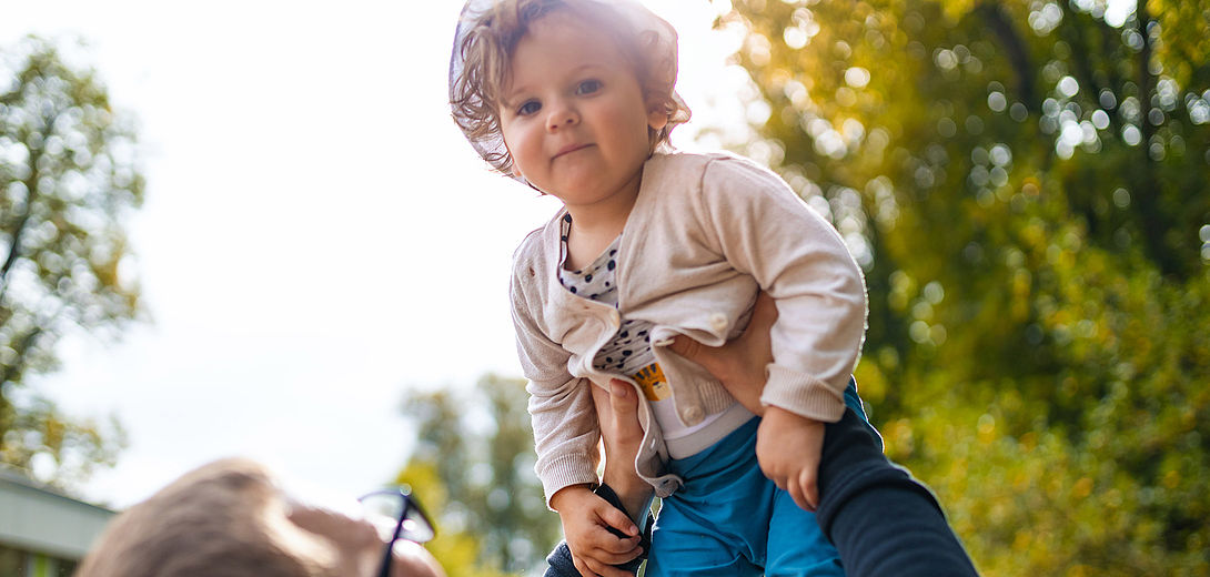Ein Kleinkind wird von einem männlichen Erzieher hochgehalten. Das Kind schaut direkt in die Kamera. Im Hintergrund sind Bäume und die Sonne im Gegenlicht.