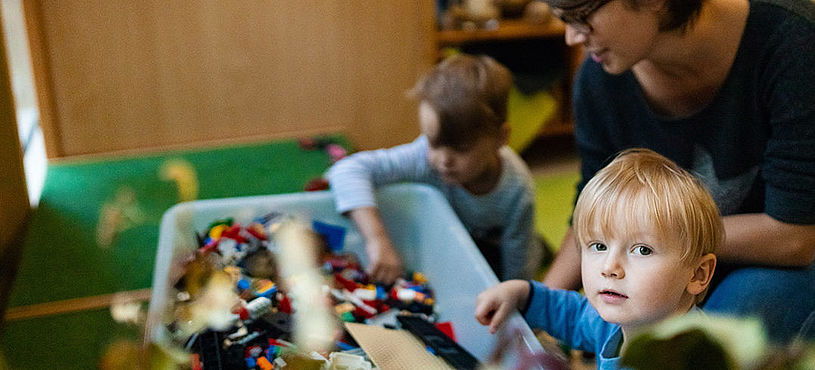 Im Vordergrund schaut ein Kind frontal in die Kamera. Im Hintergrund ein weiteres Kind Lego spielend. Eine Erzieherin kniet hinter den beiden lächelnd.