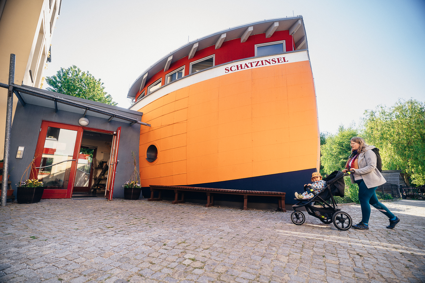 Eine Mutter mit ihrem Kind in einem Kinderwagen vor dem orangefarbenen Haus, das einem Schiff nachempfunden ist, der Kita Schatzinsel des Epilepsiezentrums Kleinwachau in Pulsnitz in Sachsen